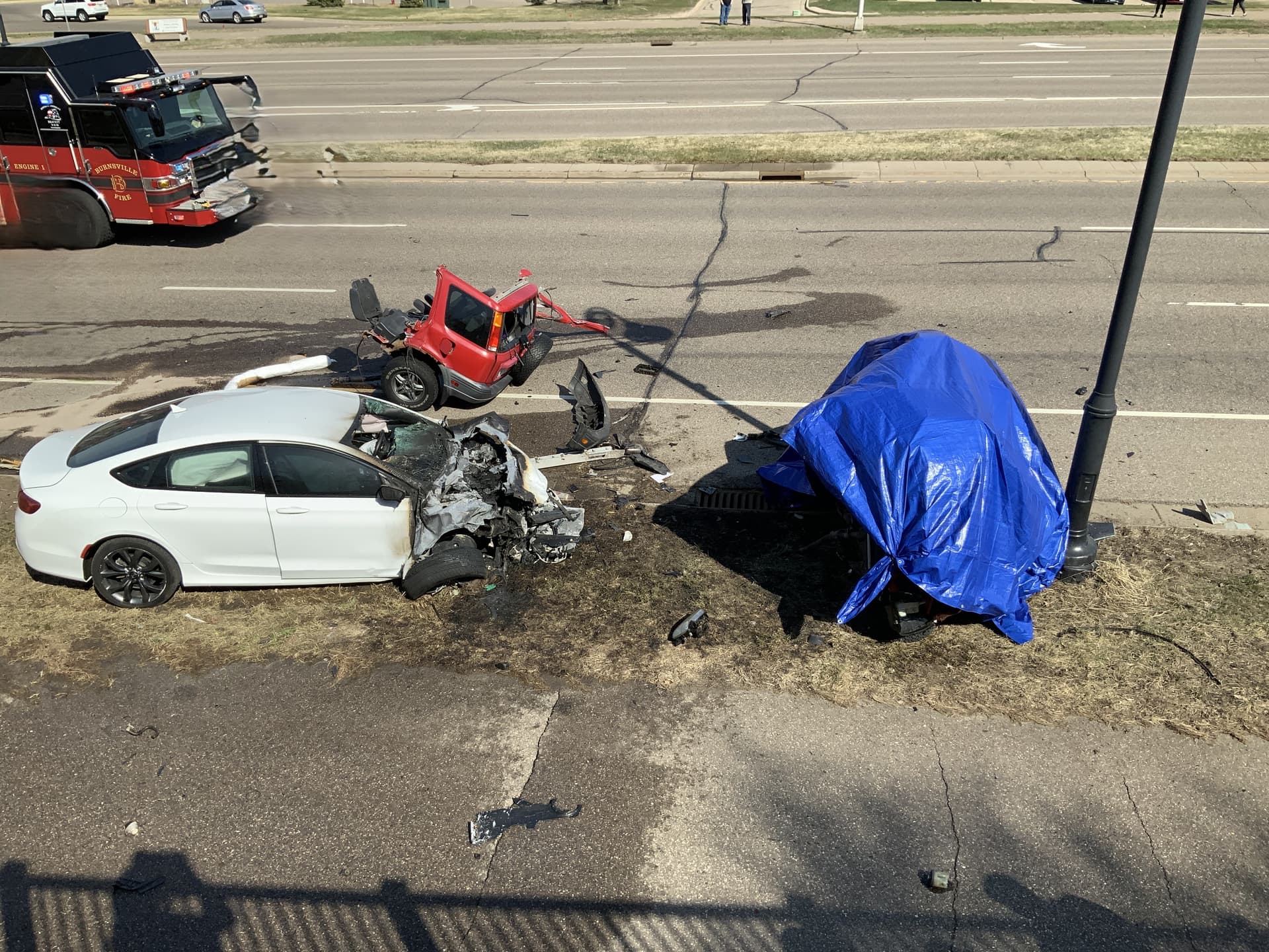 A white car with its front end missing and a piece of a red car in the street next two it. A tarp covers the rest of the crash.