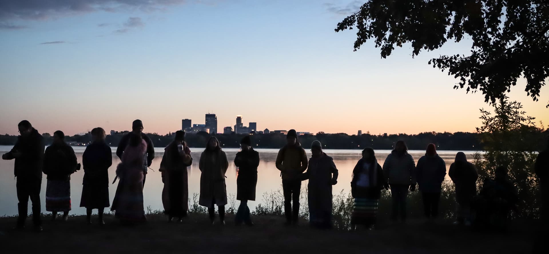 A group stands along the Mississippi River at sunrise to celebrate Indigenous Peoples Day